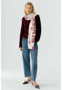 Long écharpe rose avec un motif abstrait rouge drapée sur un pull foncé. Le modèle porte un jean bleu et des chaussures à talons bordeaux. Design simple et moderne.