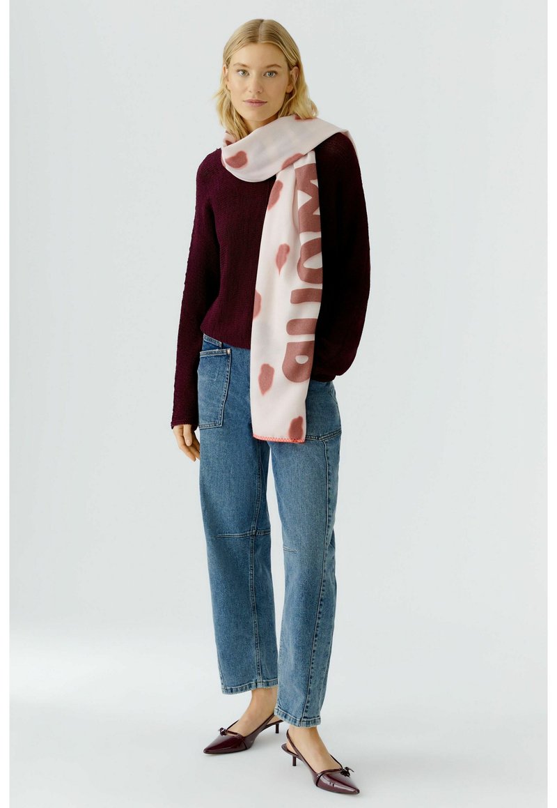 Long écharpe rose avec un motif abstrait rouge drapée sur un pull foncé. Le modèle porte un jean bleu et des chaussures à talons bordeaux. Design simple et moderne.