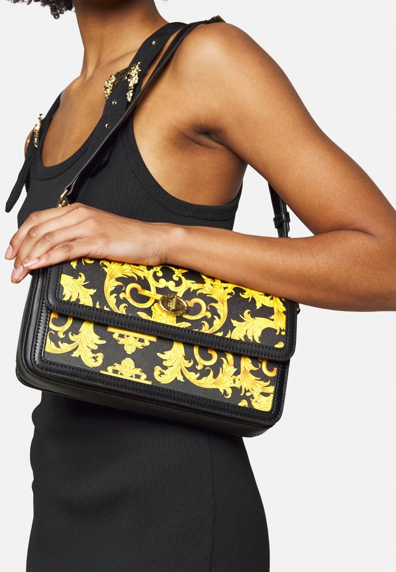 Czarna torebka z odważnym złotym wzorem w stylu barokowym, prostokątna, z klapką i złotymi dodatkami, noszona na ramieniu.