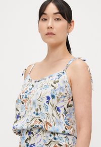 Φλοράλ τοπ με τιράντες σε ανοιχτό μπλε ύφασμα με διάφορα λουλούδια. Διαθέτει ρυθμιζόμενες δέστρες στις τιράντες και άνετη εφαρμογή με σούρες στο τελείωμα.