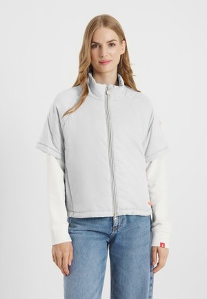 Vrouw met lang blond haar die een lichtgrijze korte mouwen jas met rits draagt over een wit shirt met lange mouwen en blauwe jeans.