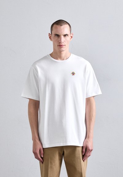 T-shirt en coton blanc à manches courtes, présentant un petit logo brodé multicolore sur la poitrine. Le modèle porte un pantalon beige.