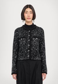 Cardigan nero con paillettes argentate, scollatura tonda, chiusura con bottoni, due tasche sul petto e polsini a coste. Indossato sopra un dolcevita nero e pantaloni a righe.