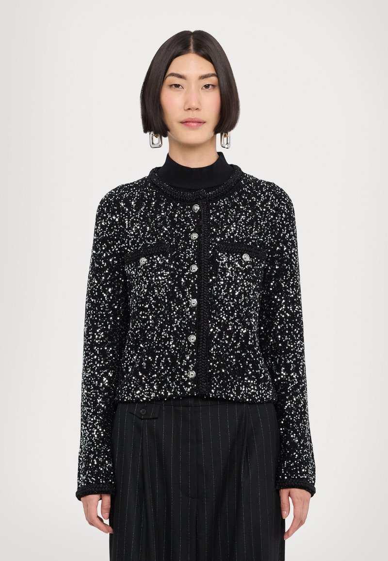 Cardigan nero con paillettes argentate, scollatura tonda, chiusura con bottoni, due tasche sul petto e polsini a coste. Indossato sopra un dolcevita nero e pantaloni a righe.
