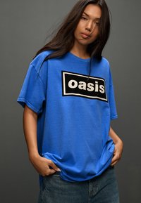 Blauw katoenen T-shirt met een losse pasvorm en korte mouwen, met een zwart rechthoek en witte tekst "oasis" aan de voorkant.