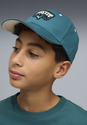 Młoda osoba nosząca morski zielony daszek z białym spodem oraz dopasowaną morskozieloną koszulkę, spoglądająca w dół i na bok na tle szarego tła.