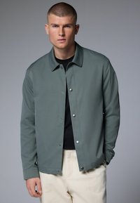 Chaqueta ligera gris con cuello tipo solapa, cierre de botones a presión y tejido texturizado. Lleva puesta sobre una camisa negra y pantalones color crema.