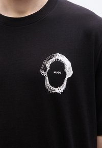 Μαύρο βαμβακερό μπλουζάκι με λευκό γραφικό στόμα καρχαρία και το κείμενο "HUGO" μέσα. Κοντά μανίκια και λαιμόκοψη τύπου crew.