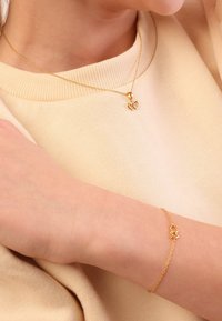 Gouden ketting met twee verstrengelde hartbedels en een bijpassende delicate armband, gedragen over een beige trui. Glad, glanzend metalen afwerking.