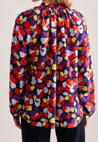 Blommig blus med ett flerfärgat mönster i rött, lila, gult och blått. Långa ärmar med rynkad halsringning och en luftig passform.