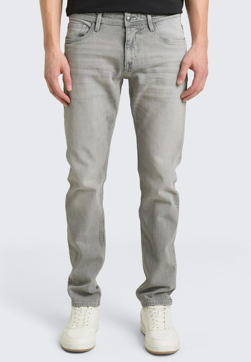 Persona che indossa jeans aderenti grigio chiaro e sneakers bianche, in piedi contro uno sfondo chiaro e semplice.
