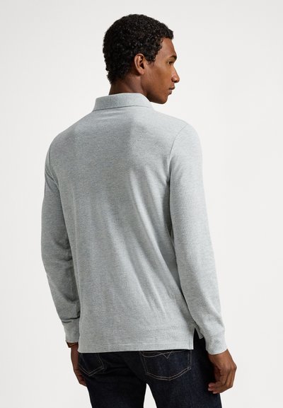 Ανοιχτό γκρι πόλο με μακρύ μανίκι και γιακά, υφασμένο με υφή και πλευρικές σχισμές, σε συνδυασμό με σκούρο τζιν. Πλάγια όψη από πίσω, που δείχνει τη εφαρμογή και το στυλ.