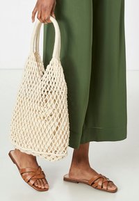 Woven straw tote bag in cream with two sturdy handles, paired with brown leather slide sandals. Background features green fabric.