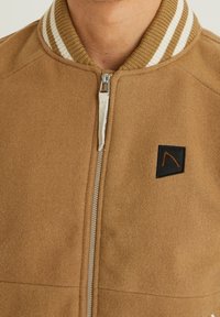 Brun ullblandning bomberjacka med dragkedja fram. Har en ribbstickad krage med bruna och vita ränder samt en svart logotyp patch.