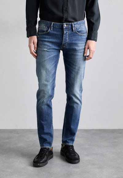 Jeans de denim pentru bărbați, de culoare albastră, cu o croială slim, având un design estompat și ușor deteriorat. Asortați cu adidași negri pe un fundal neutru.