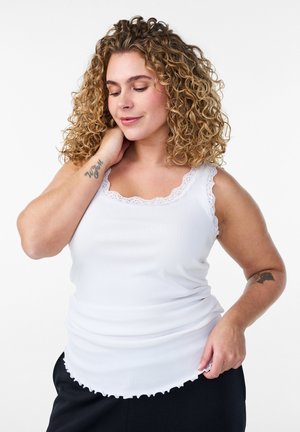 Curly-haired woman in white sleeveless top with lace trim touches neck and holds hem, showing wrist tattoos, against plain white background.