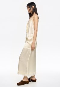 Camisole en satin crème avec des ornements en dentelle, associée à une longue jupe assortie. L'ensemble présente des bretelles fines et une coupe décontractée. Des mules marron viennent compléter le look.