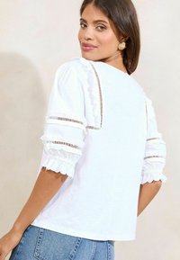 Hvid bluse med korte, rynkede ærmer. Har blonde detaljer langs sømmene og en afslappet pasform, parret med blå denim jeans.