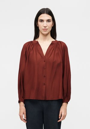 Frau mit langen dunklen Haaren trägt eine lockere, geknöpfte, rostfarbene Bluse mit gerafften Schultern und schwarze Hosen, steht vor weißem Hintergrund.