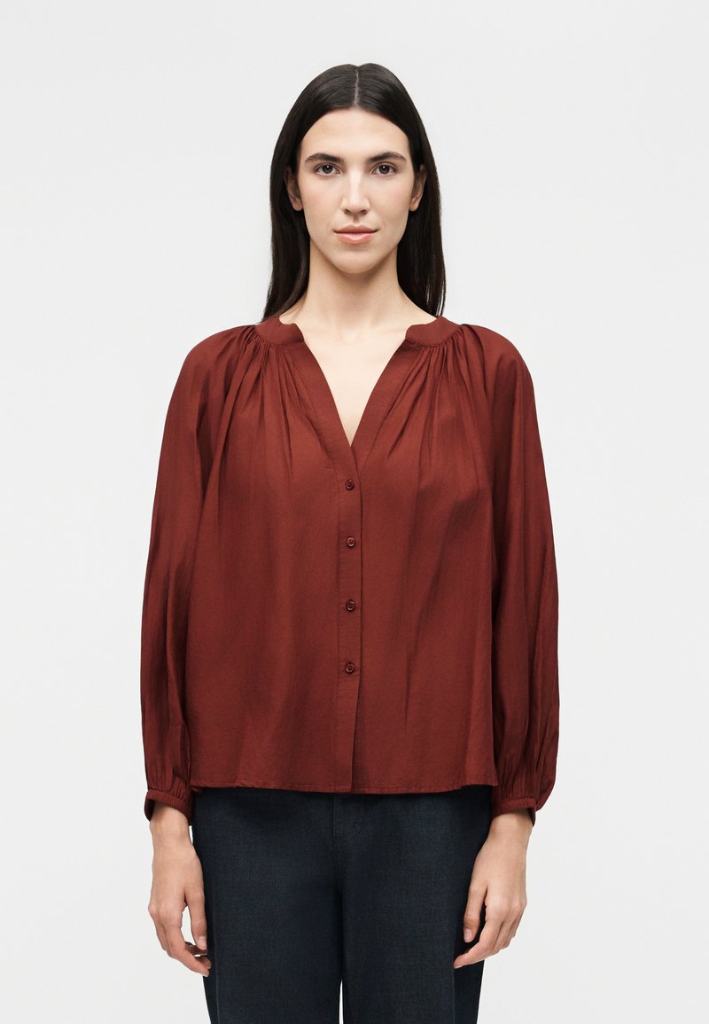 Γυναίκα με μακριά σκούρα μαλλιά φοράει μια φαρδιά, κουμπωτή, σκουροκόκκινη μπλούζα με σούρες στους ώμους και μαύρο παντελόνι, στεκόμενη μπροστά σε λευκό φόντο.