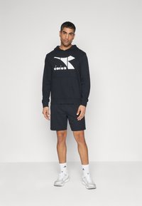 Felpa nera con logo "diadora" bianco, tasca a marsupio, abbinata a pantaloni corti neri e scarpe da ginnastica bianche. Abbigliamento sportivo semplice e informale.