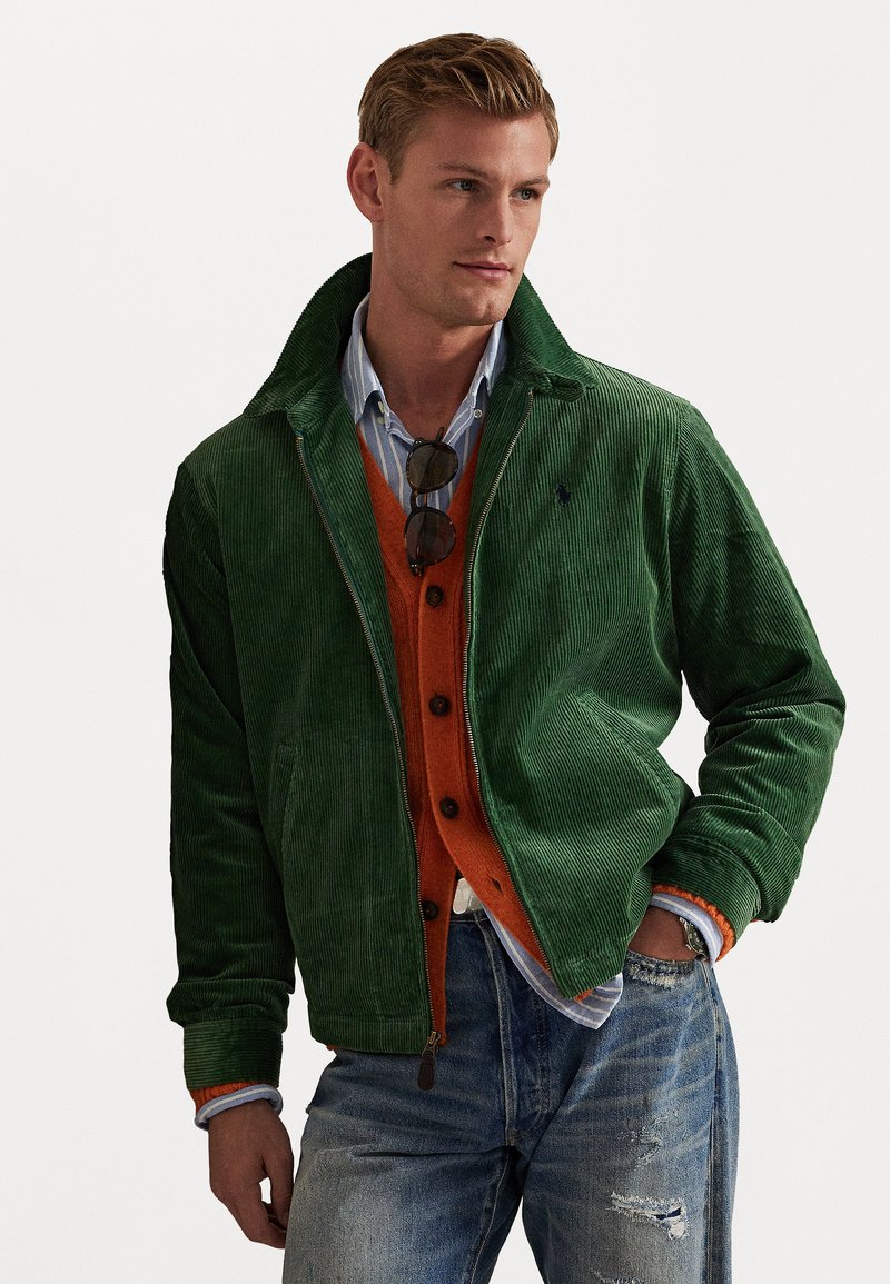 Giacca in corduroy verde con zip frontale, dotata di colletto, tasche laterali e sottili costine; indossata sopra un cardigan arancione e una camicia blu.