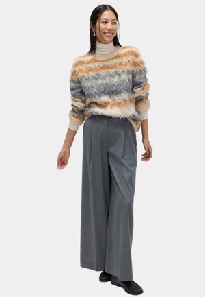 Pull en maille texturée à rayures orange, grises et crème ; col montant. Pantalon gris à jambes larges ; chaussures plates noires. Boucles d'oreilles simples.