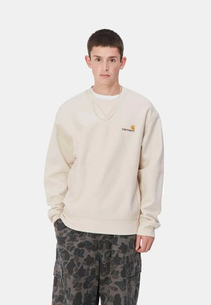 Sudadera beige con puños y dobladillo acanalados, que presenta un pequeño logo dorado. Llevada sobre una camisa blanca y combinada con pantalones cargo de camuflaje.
