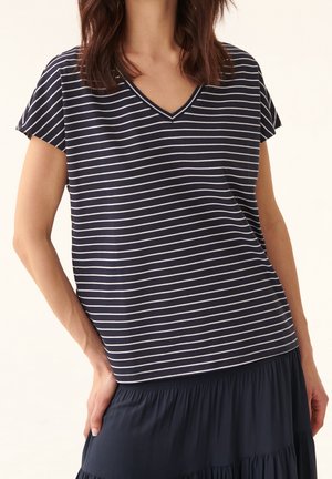 Vrouw draagt een marineblauw-witte horizontaal gestreepte V-hals top met korte mouwen en een marineblauwe rok, hand rustend op de heup.