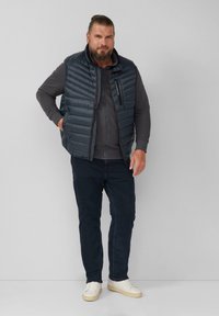 Dunkelblaue, gepolsterte Weste mit horizontalem Steppmuster, getragen über einem grauen Sweatshirt mit Reißverschluss. Kombiniert mit dunklen Jeans und weißen Sneakern.