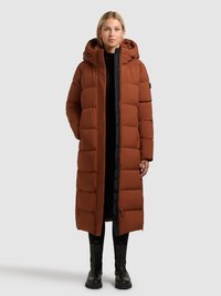 Dlouhý hnědý péřový kabát s kapucí, prošívaným designem, černými zipy a bočními kapsami. Nošený přes černý outfit.