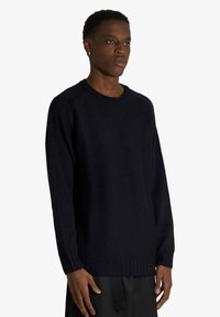 Giovane uomo che indossa un maglione nero semplice a maglia e pantaloni neri, in piedi davanti a uno sfondo bianco, rivolto leggermente verso destra.