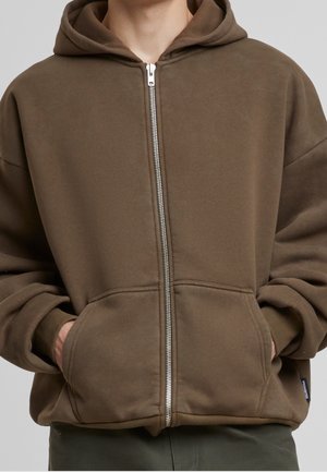 Osoba nosząca brązową bluzę z kapturem zapinaną na zamek, z rękami w przednich kieszeniach, widocznym zamkiem błyskawicznym i ściągaczami na rękawach.