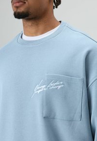 Lichtblauwe sweater gemaakt van katoen met een ronde hals, voorzien van een voorzak en witte geborduurde tekstdetails. Zachte textuur.