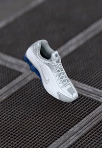 White and gray Nike running shoe with blue sole detail, laced up, positioned on industrial metal grid background.