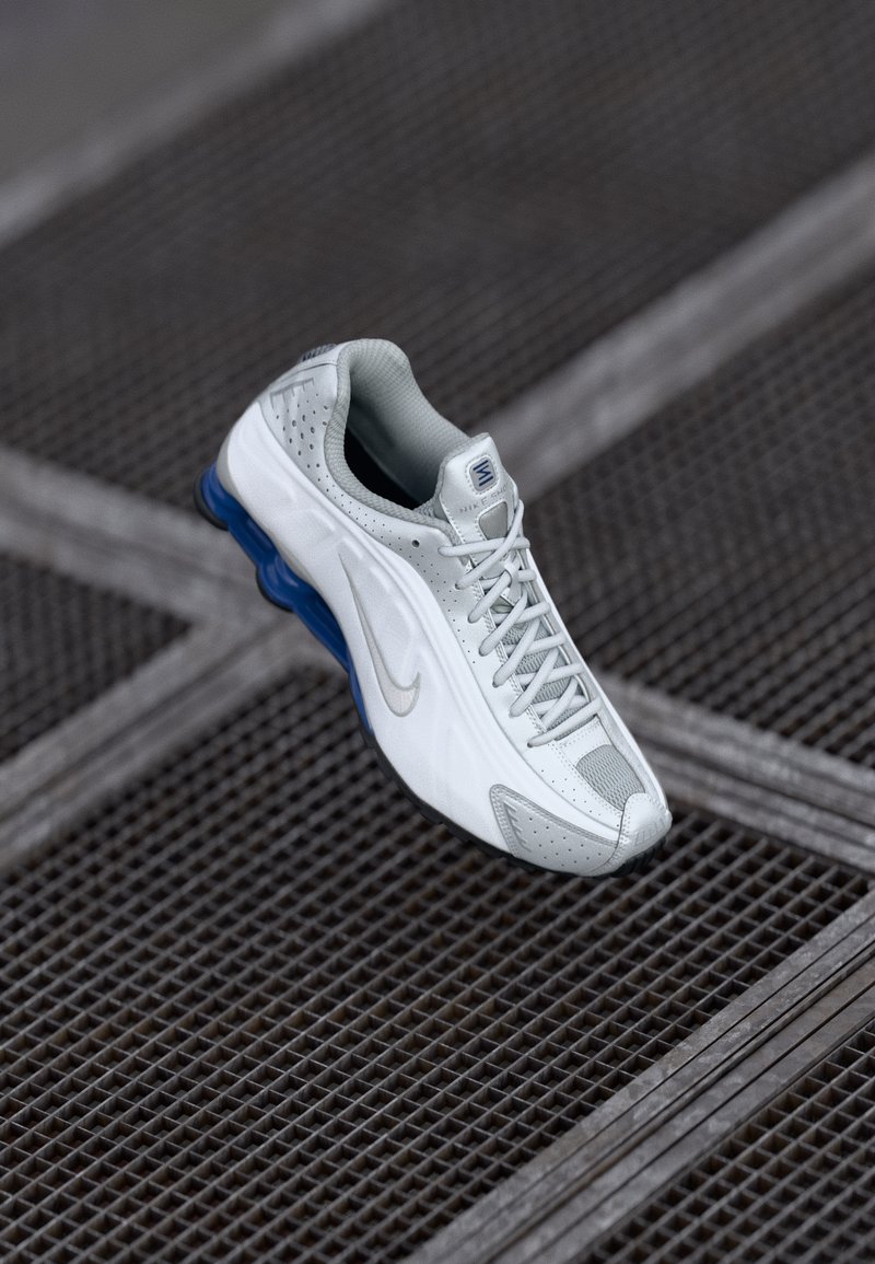 White and gray Nike running shoe with blue sole detail, laced up, positioned on industrial metal grid background.