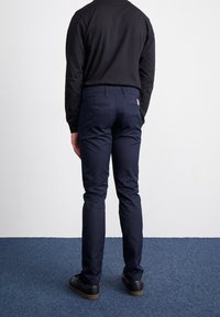 Marineblaue Baumwollhose mit geradem Schnitt, ausgestattet mit Gesäßtaschen und einem dezenten Logo-Tag, kombiniert mit schwarzen Schuhen vor einem blauen Teppichhintergrund.