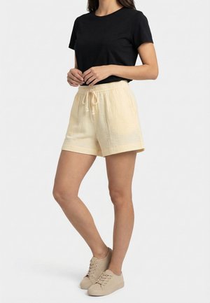 Mujer que lleva una camiseta negra de manga corta, pantalones cortos beige con cordón y zapatillas beige, de pie con las manos juntas delante.