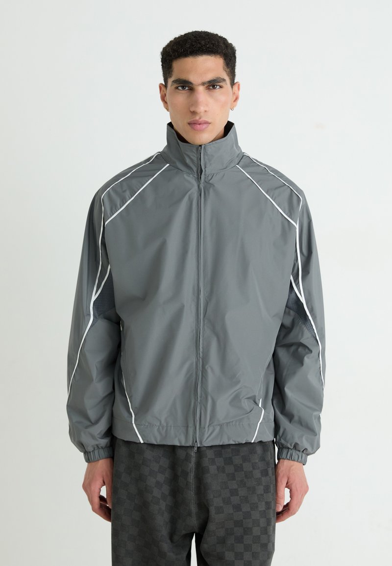 Veste coupe-vent grise avec col haut, accents blancs et poignets élastiques ; présente des panneaux latéraux texturés pour la ventilation.