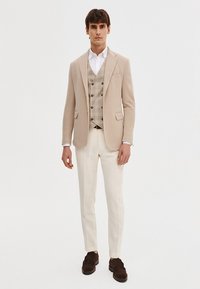 Beżowa marynarka o dopasowanym kroju, noszona na wiskozy w kratę, zestawiona z jasnymi spodniami. Brązowe zamszowe buty i biała koszula dopełniają całość.
