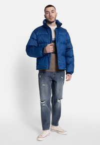 Modrá páperová bunda s vysokým golierom, v kombinácii s pleteným sveterom so svetlým vzorom a roztrhnutými modrými džínsy.