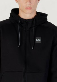 Schwarze Zip-Hoodie aus weichem Stoff, mit Kapuze mit Kordelzug, Fronttaschen und einem kleinen EA7-Logo-Patch auf der Brust.
