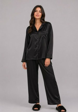 Femme debout vêtue d’un ensemble pyjama en satin noir à manches longues et pantalon, assorti de chaussons noirs duveteux, sur un fond gris uni.