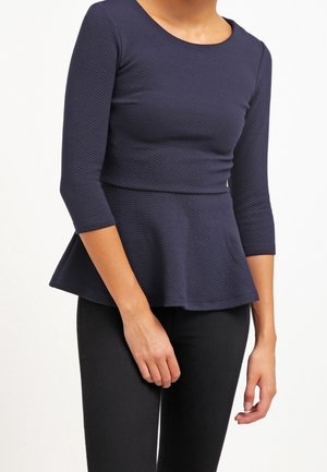 Kobieta ubrana w dopasowany granatowy top peplum z rękawami do łokcia i czarne spodnie, stojąca na jednolitym tle.
