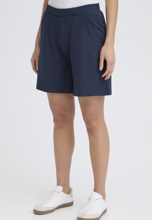 Osoba ubrana w granatowe krótkie spodenki do kolan z bocznymi kieszeniami oraz białe trampki na tle jednolitym.