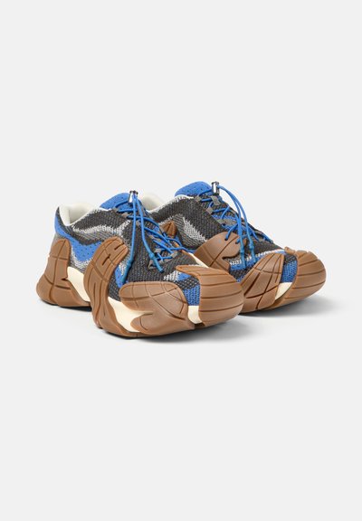 Zapatillas con suela gruesa en tejido azul, gris y blanco con superposiciones de goma marrón y cordones con cuerda azul, colocadas en ángulo una al lado de la otra sobre un fondo blanco.