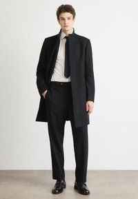 Palton negru din lână cu guler înalt, purtat peste o cămașă albă cu guler și o cravată neagră, asortat cu pantaloni negri la comandă și pantofi negri eleganți.