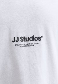 Valge puuvillane t-särk, millel on must logotekst "JJ Studios" lisatekstiga allpool, sileda ja ühtlase kangaga.