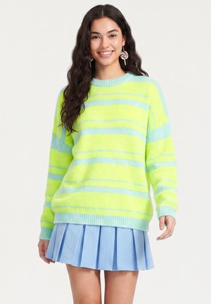 Ung kvinde med langt, mørkt hår iført en gul og lyseblå stribet sweater, lyseblåt plisseret skørt og farverige slikkepind-øreringe, der smiler.