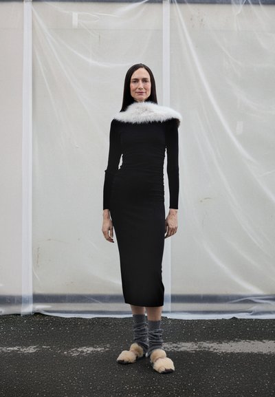 Fekete, testhez simuló ruha hosszú ujjakkal, fehér műszőrme gallérral díszítve. Szürke mintás zoknival és bézs, bolyhos papuccsal viselve. Egyszínű háttér.